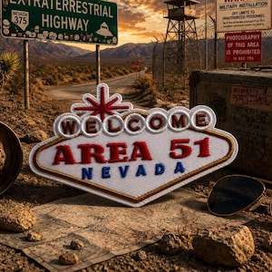 Pode incluir: Um patch com as palavras "WELCOME AREA 51 NEVADA" em vermelho e azul, semelhante ao icónico letreiro de Las Vegas. A imagem inclui um sinal de estrada para a Extraterrestrial Highway, um mapa, óculos de sol e uma instalação militar.