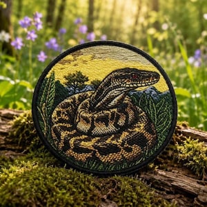 May include: A circular embroidered patch featuring a detailed brown and black snake coiled in a grassy landscape. The background depicts a yellow sky and green trees, all enclosed within a solid black stitched border edge.