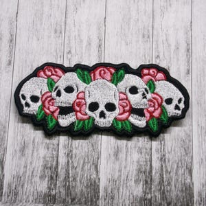 May include: Embroidered patch featuring five white skulls with black eye sockets and pink roses. The skulls are surrounded by green leaves and the patch is black.