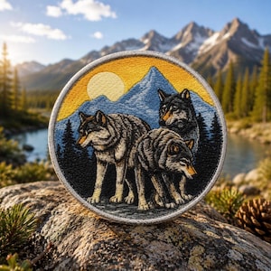 May include: Embroidered patch featuring three wolves in a mountain landscape with a yellow sun and blue mountains. The patch is circular with a detailed design and a stitched border. The background shows a river and trees.