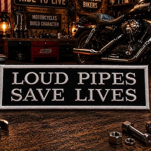 May include: A rectangular black embroidered patch with a white border and the white stitched text LOUD PIPES SAVE LIVES. The patch features a clean, bold font and is presented in a horizontal orientation, showcasing high-contrast lettering.