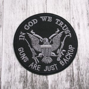 May include: Black and white embroidered patch with a bald eagle and the text "In God We Trust Guns Are Just Backup".