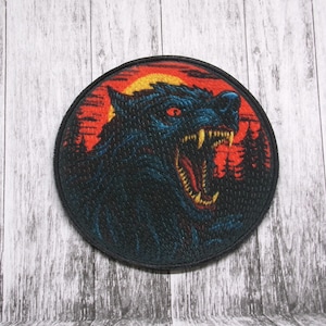 Może przedstawiać: Okrągła naszywka przedstawiająca warczącego wilka z czerwonymi oczami i ostrymi zębami na tle zachodu słońca. Wilk jest głównie czarny i niebieski, z czerwonym i żółtym niebem oraz sylwetką lasu.