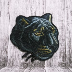 Pode incluir: Patch bordado com a cabeça de uma pantera negra, com um design detalhado e olhos amarelos. O patch está sobre um fundo de madeira.