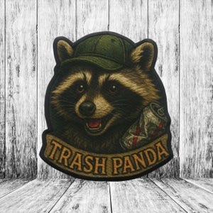 Könnte beinhalten: Gestickter Aufnäher mit einem Waschbären, der eine grüne Kappe trägt und eine zerdrückte Dose hält. Der Aufnäher trägt die Aufschrift "Trash Panda" auf einem Banner. Der Waschbär hat braunes und schwarzes Fell.