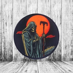 May include: A black and blue embroidered patch featuring a skeletal figure in a hooded robe holding a staff, standing in front of a large orange sun.