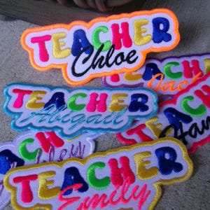 May include: Embroidered patches with the word "TEACHER" in colorful letters, each with a different name below. The patches have a white border and are in various bright colors, including pink, blue, yellow, and green.