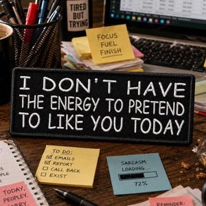 May include: A rectangular black embroidered patch featuring white stitched text that reads I DON'T HAVE THE ENERGY TO PRETEND TO LIKE YOU TODAY. The patch has a clean, finished border and is positioned horizontally on a dark wooden desk surface.