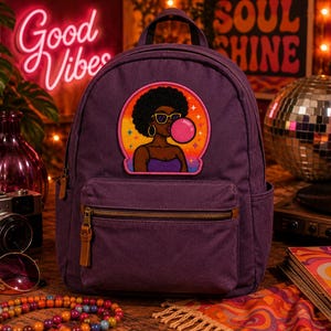 Pode incluir: Uma mochila roxa escura com um emblema bordado central de uma pessoa com penteado afro, usando &oacute;culos de sol e fazendo uma bolha rosa. A mochila possui um bolso frontal com z&iacute;per, puxadores de couro castanho e bolsos laterais.