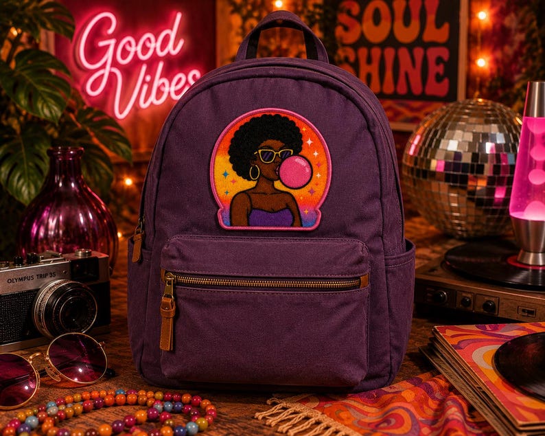 Pode incluir: Uma mochila roxa escura com um emblema bordado central de uma pessoa com penteado afro, usando &oacute;culos de sol e fazendo uma bolha rosa. A mochila possui um bolso frontal com z&iacute;per, puxadores de couro castanho e bolsos laterais.