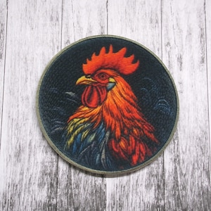 May include: A round embroidered patch featuring a colorful rooster with a red comb and wattle. The rooster is depicted against a dark background with a green and blue feathered design.
