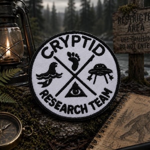 May include: A circular embroidered patch with a white background and black border. The text "CRYPTID RESEARCH TEAM" is stitched in black. The center features a black cross design with icons of a sea creature, a footprint, a UFO, and an eye.