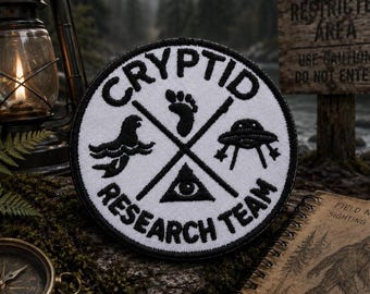 May include: A circular embroidered patch with a white background and black border. The text "CRYPTID RESEARCH TEAM" is stitched in black. The center features a black cross design with icons of a sea creature, a footprint, a UFO, and an eye.