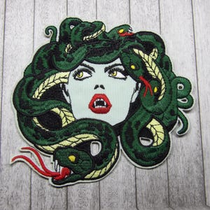 May include: Embroidered patch depicting a woman with green hair made of snakes. The woman has a fierce expression with fangs showing. The patch is ideal for adding a touch of Greek mythology to your clothing or accessories.