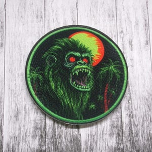 May include: Round patch featuring a green ape with red eyes, baring its teeth. A green and orange sun is in the background, with palm trees on either side. The patch has a black border.