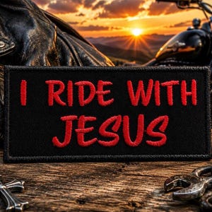 May include: Black rectangular patch with the red text "I RIDE WITH JESUS". The patch is on a wooden surface with a leather jacket, motorcycle, and cross keychain in the background. The sunset is visible in the background.