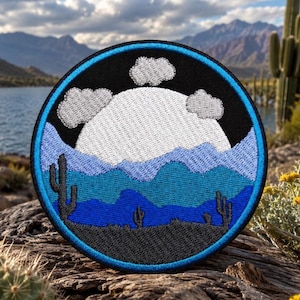 May include: A circular embroidered patch featuring a desert landscape with a large white moon, grey clouds, and layered blue mountains. The foreground shows dark grey silhouettes of saguaro cacti against a black sky, all bordered by a bright blue rim.