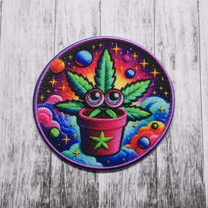 May include: A colorful embroidered patch featuring a cartoon marijuana plant with eyes in a pink pot. The plant is surrounded by a psychedelic background of stars, planets, and clouds.