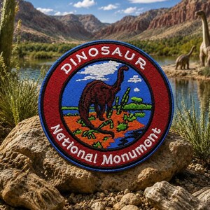Może przedstawiać: Okrągła naszywka z napisem "DINOSAUR National Monument" w białych literach na czerwonym i niebieskim tle. Naszywka przedstawia brązową ilustrację dinozaura. Naszywka jest umieszczona na tle malowniczych gór i wody.
