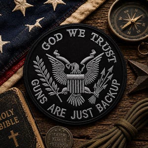 May include: A black patch with the text "GOD WE TRUST GUNS ARE JUST BACKUP" and an eagle emblem. The patch is surrounded by an American flag, a compass, a Bible, and other patriotic items on a wooden surface.