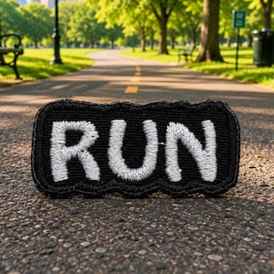 May include: A rectangular embroidered patch with a black background and the word RUN stitched in white thread. The patch features a thick, textured border and a bold, sans-serif font style, creating a clear contrast between the white letters and black fabric.