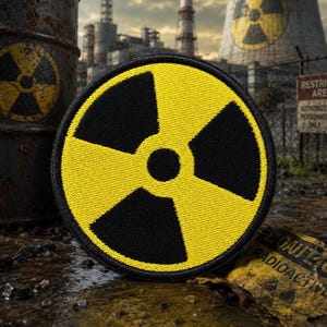 May include: A circular embroidered patch featuring a black and yellow radiation hazard symbol. The design consists of a central black circle surrounded by three black blades separated by yellow sections, all enclosed within a black stitched border.