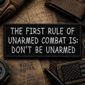 May include: A rectangular black morale patch with a stitched border featuring the white embroidered text: THE FIRST RULE OF UNARMED COMBAT IS: DON'T BE UNARMED. The patch has a clean, bold font and a durable fabric construction with a finished edge.
