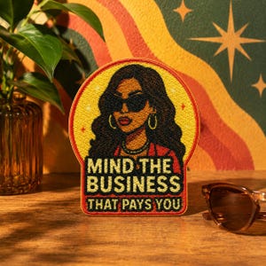 May include: Embroidered patch featuring a stylish woman in sunglasses, hoop earrings, and a red jacket. The patch is circular with a yellow background and the text "MIND THE BUSINESS THAT PAYS YOU" in black and white. The patch is on a wooden surface.