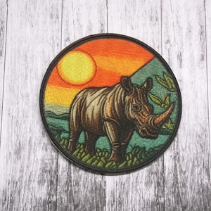 Pode incluir: Um remendo redondo bordado com um rinoceronte em pé em um campo verde com um pôr do sol ao fundo. O sol é amarelo e o céu é laranja e vermelho.