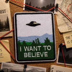 May include: A rectangular embroidered patch featuring a black UFO hovering over a landscape of green pine trees and light blue mountains. The text I WANT TO BELIEVE is stitched in white capital letters across the bottom of the patch.
