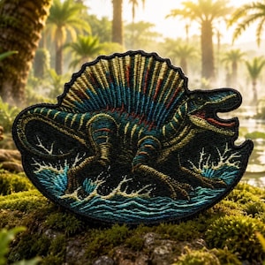 Könnte beinhalten: Ein gestickter Aufnäher mit einem Spinosaurus-Dinosaurier mit einem markanten Rückensegel. Der Dinosaurier ist in Dunkelgrün, Blau und Gold gehalten und steht in blauem Wasser mit weißen Spritzern. Der Aufnäher hat einen schwarzen Rand.