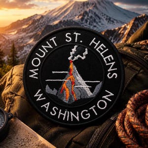 Może przedstawiać: Okrągła czarna naszywka z białym haftowanym napisem „Mount St. Helens Washington”. Naszywka przedstawia ilustrację wulkanu w erupcji z pomarańczowymi i szarymi detalami. Tło przedstawia pasmo górskie.