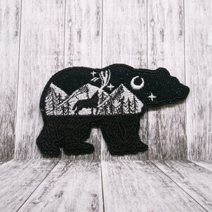 May include: Black bear-shaped patch with an embroidered mountain scene. The patch features a white mountain range, trees, a deer, a crescent moon, and stars. The bear's outline has a glittery finish.