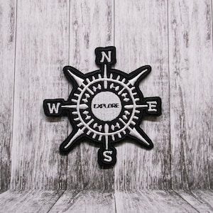 Puede incluir: Parche de brújula en blanco y negro con la palabra "Explore" en el centro y las letras N, S, E y W alrededor de la rosa de los vientos.