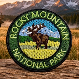 May include: A circular embroidered patch featuring a brown elk standing in a yellow field with a blue mountain range in the background. The border is dark green with the text "ROCKY MOUNTAIN NATIONAL PARK" stitched in light green lettering.