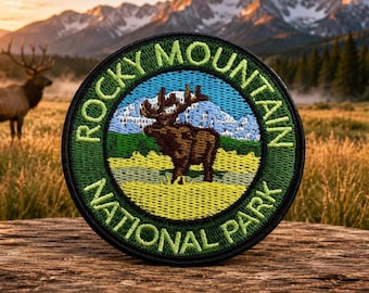 May include: A circular embroidered patch featuring a brown elk standing in a yellow field with a blue mountain range in the background. The border is dark green with the text "ROCKY MOUNTAIN NATIONAL PARK" stitched in light green lettering.