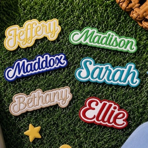 Puede incluir: Seis parches de nombre personalizados en amarillo, verde, azul, verde azulado, beige y rojo con letras blancas. Los nombres Jeffery, Madison, Maddox, Sarah, Bethany y Ellie se muestran en una fuente cursiva negrita sobre una superficie verde.
