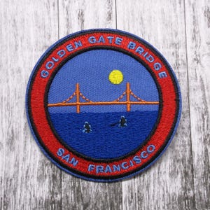 May include: A red, white, and blue embroidered patch with the words "Golden Gate Bridge San Francisco" around the edge. The center of the patch features an orange embroidered image of the Golden Gate Bridge with a yellow sun in the sky.