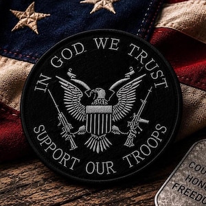 May include: A circular black embroidered patch featuring a silver eagle emblem with a shield, flanked by two rifles. The text IN GOD WE TRUST and SUPPORT OUR TROOPS is stitched in silver thread around the border of the circular patch design.