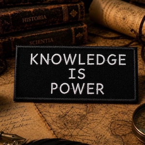 Peut inclure: Un écusson rectangulaire noir avec l'inscription blanche brodée « KNOWLEDGE IS POWER ». L'écusson est posé sur une carte vintage, entouré de livres anciens, d'une bougie et d'une loupe.