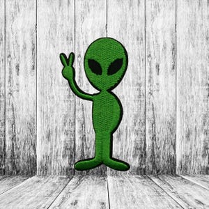 May include: Green alien patch with black eyes and outline, giving a peace sign. The embroidered patch is on a white and gray wood grain background. The alien is standing and has a peace sign hand gesture.