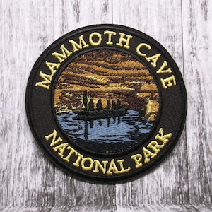 Puede incluir: Un parche redondo negro con letras doradas y un diseño de paisaje detallado. El texto dice "MAMMOTH CAVE NATIONAL PARK". La imagen representa un barco en un lago azul con un fondo marrón y dorado, probablemente una puesta o salida de sol.
