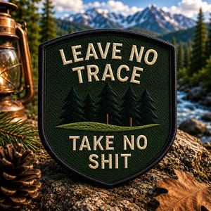 Op de afbeelding: Een donkergroene patch met de woorden "LEAVE NO TRACE" en "TAKE NO SHIT" geborduurd in wit. De patch toont een boslandschap met dennenbomen. De achtergrond toont een bergketen, een rivier en een brandende lantaarn.