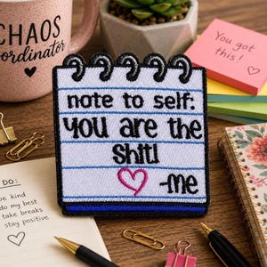 May include: A square embroidered patch designed to look like a notepad page with black spiral binding at the top. The white fabric features blue horizontal lines and black text that reads: note to self: you are the shit! -me, with a pink heart.