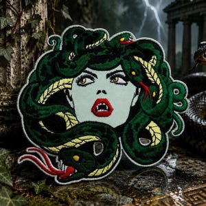 May include: Embroidered patch featuring Medusa with green snakes for hair, a pale green face, and red lips. The patch has a detailed design with yellow and red accents, perfect for jackets, bags, or other fabric items.