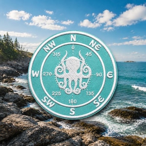May include: A round, teal patch with a white octopus compass design. The compass rose features directional markings and degree measurements. The patch is set against a scenic coastal background with rocks, ocean, and a blue sky with clouds.