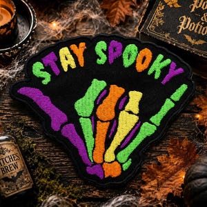 May include: A black embroidered patch featuring a skeleton hand making a shaka sign gesture in vibrant neon green, orange, yellow, and purple thread. The text STAY SPOOKY is embroidered in matching neon colors across the top of the patch.