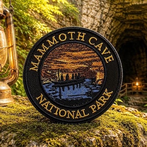 May include: A round black patch with gold lettering that reads "Mammoth Cave National Park." The patch features a landscape scene with a blue lake, a boat, and a sunset. The patch is on a mossy surface.