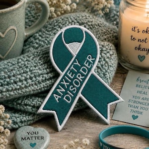 May include: A teal ribbon patch with the words "ANXIETY DISORDER" embroidered in white. A teal mug with a heart design, a candle with the text "it's okay to not be okay", and a bracelet with the text "YOU" are also visible.