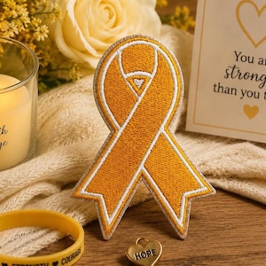 May include: An embroidered yellow ribbon patch with a white border, a gold heart charm with the word "HOPE", and a yellow rubber bracelet with the words "HOPE, STRENGTH, COURAGE". A lit candle and a card with the words "You are stronger than you think" are also present.
